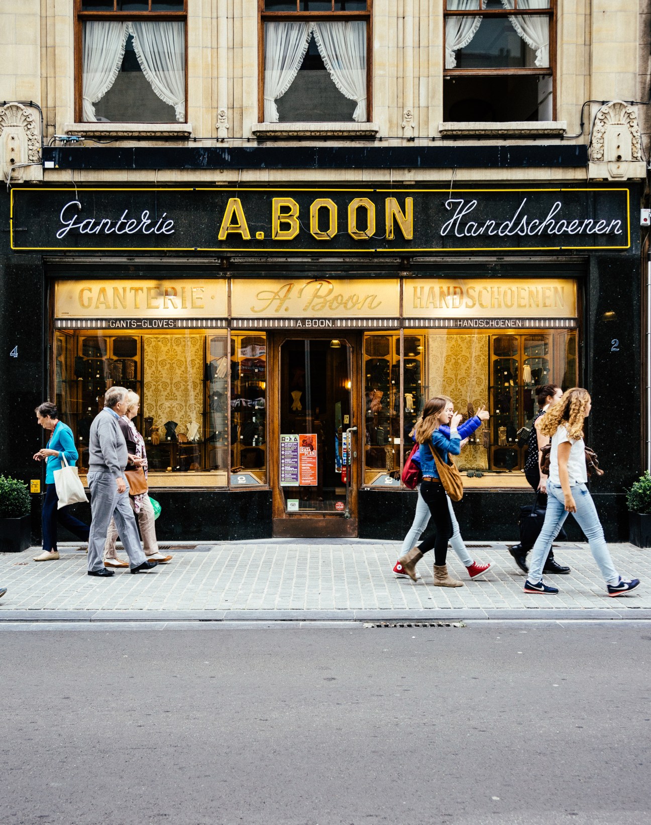 A storefront of Ganterie Boon, a historic glove shop with black facade, gold signage, and white window curtains, with pedestrians passing by on the street.