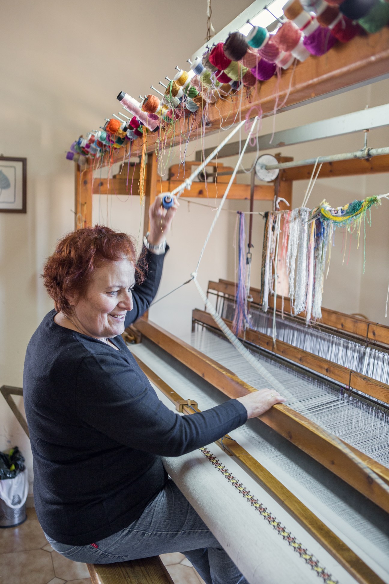 Weaver Gogo Aligizaki working at her loom with colorful yarn spools overhead.