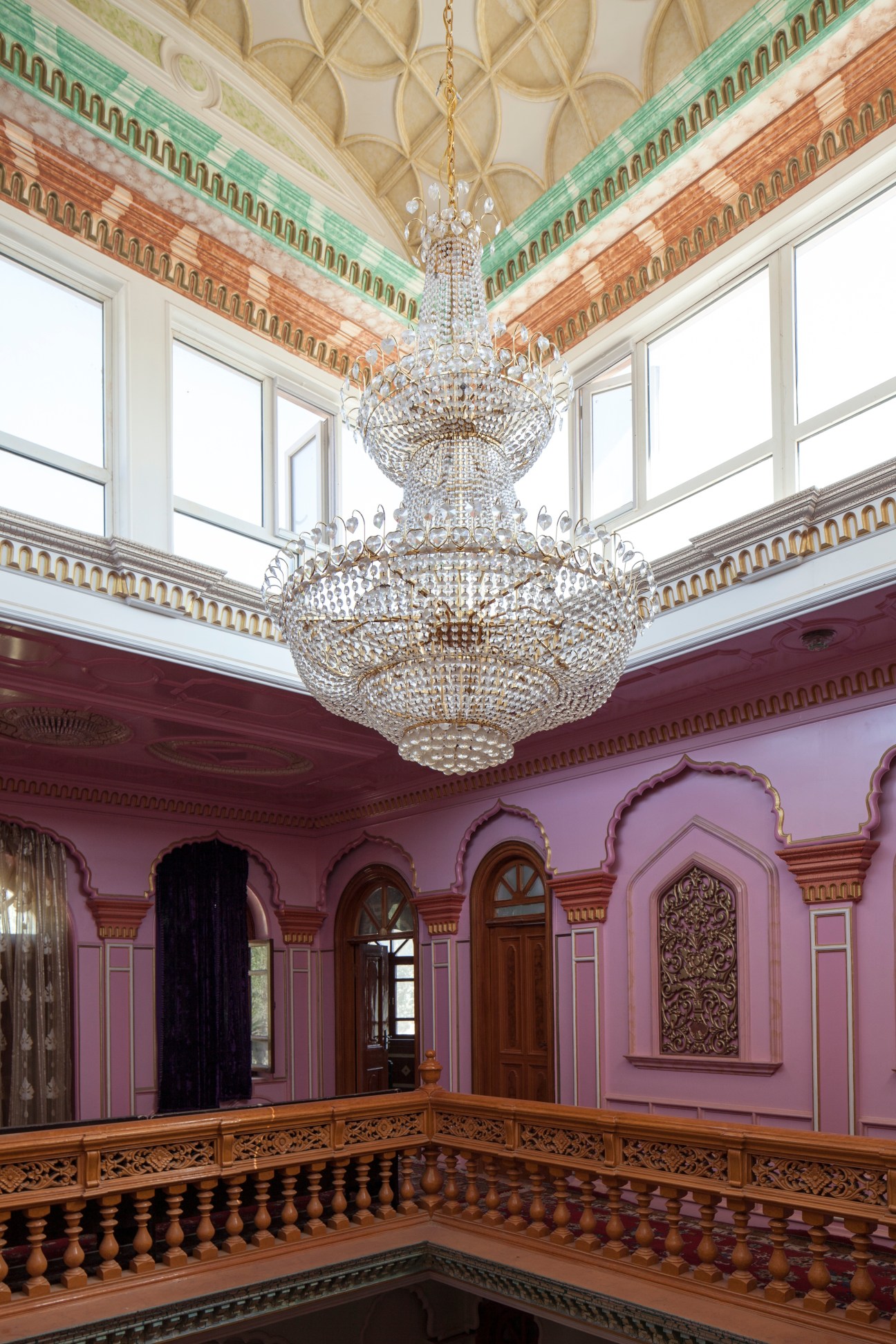 Ornate interior room with pink walls, crystal chandeliers, decorated ceiling, and wooden railings.