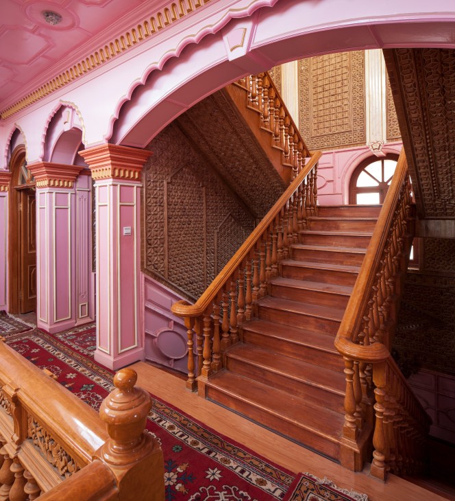 Pink-painted hallway with ornate wooden staircase and carved architectural details in Ibrahm Musa's villa.