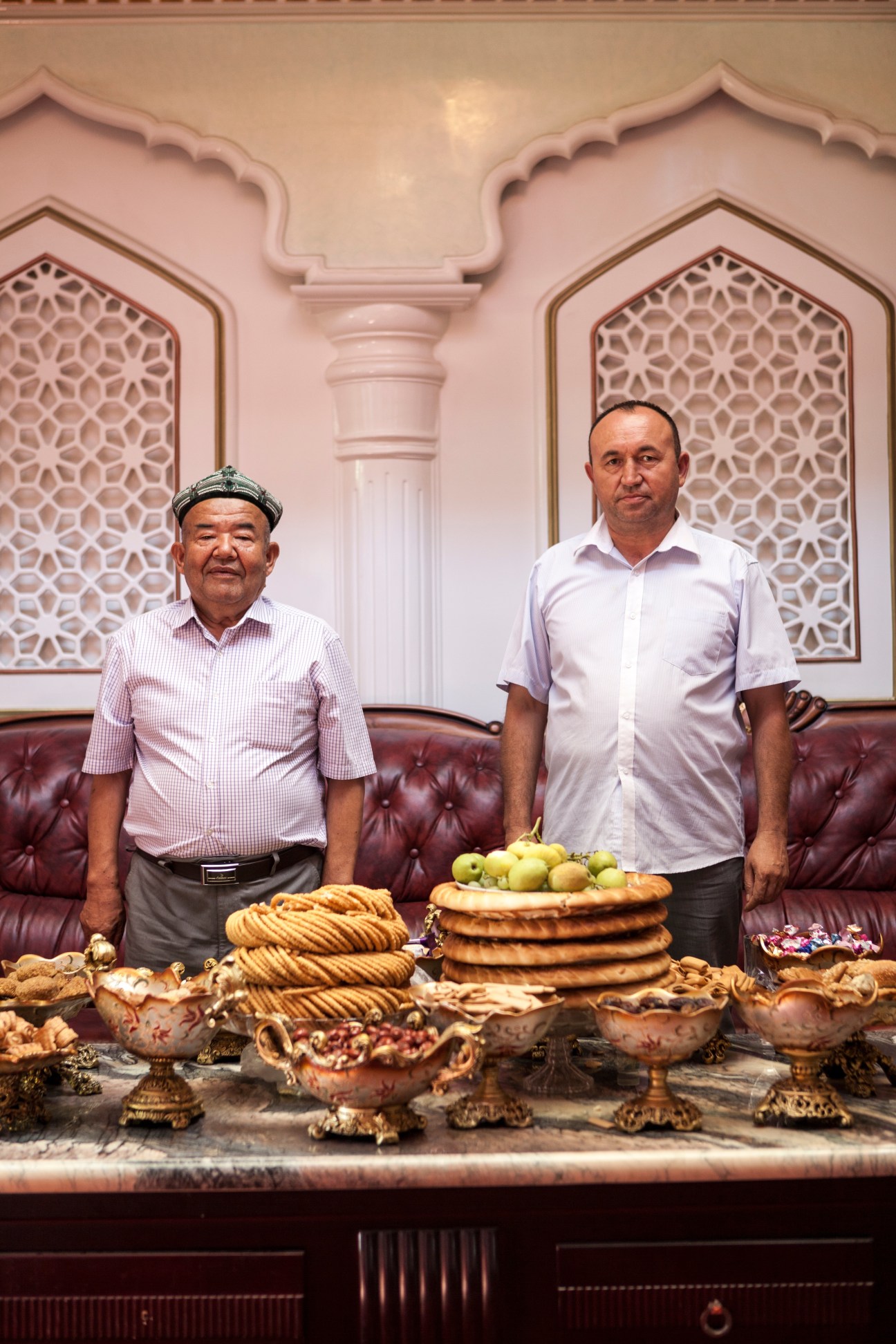 Two men stand behind a table displaying traditional Uyghur breads and pastries in an ornately decorated room with pink walls and geometric Islamic patterns.
