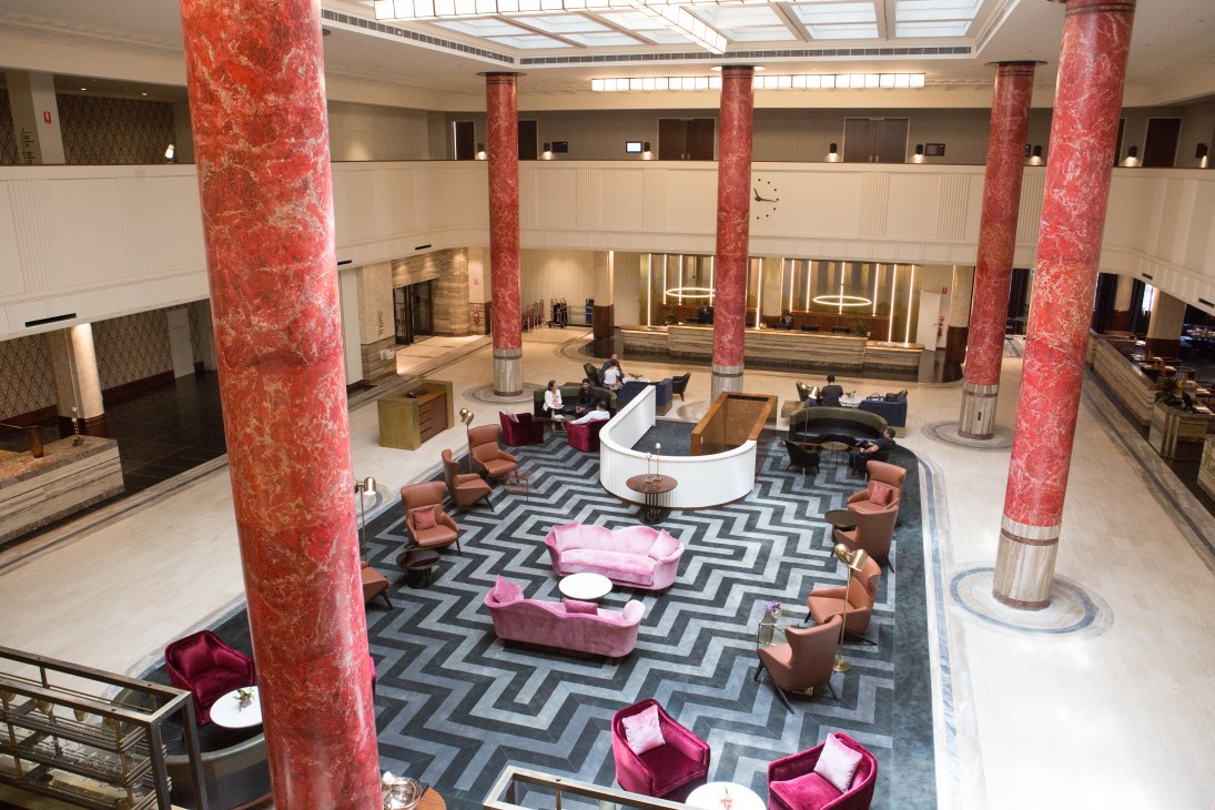 Primus Hotel's grand lobby featuring red marble pillars, art deco skylights, geometric patterned flooring, and pink velvet seating.