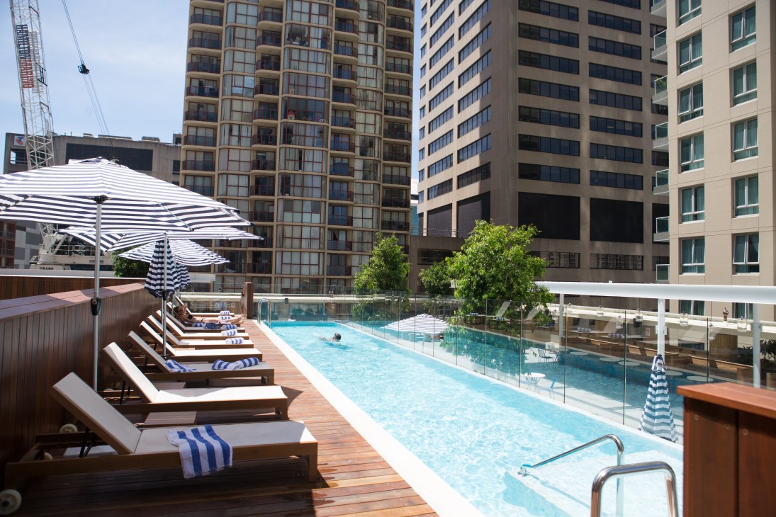 Rooftop pool at Primus Hotel with striped umbrellas, loungers and city buildings.