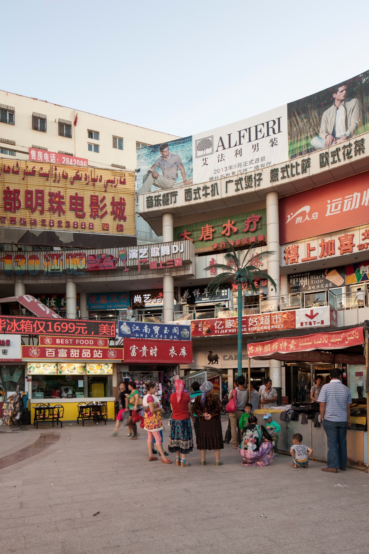 Busy outdoor shopping center with Chinese signage, storefronts, and pedestrians including women in traditional dress and children.