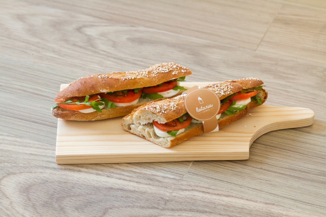 Three seeded bread sandwiches with fresh cheese, tomato and greens on a wooden board with a Leitaria Lisboa branded tag.