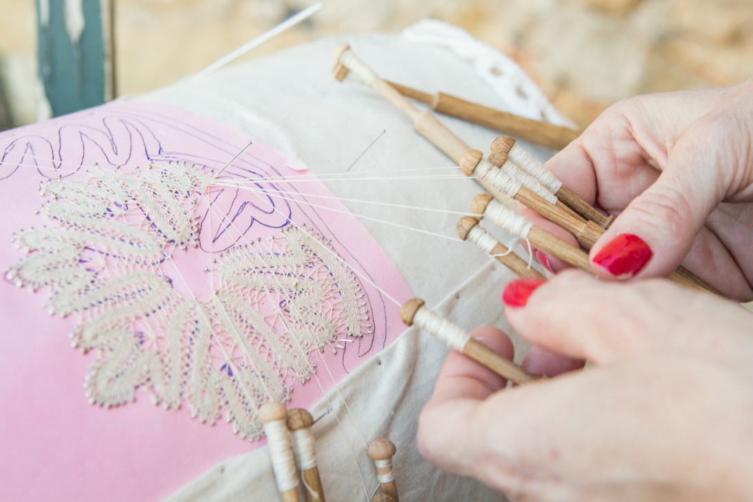 Hands with red nail polish working on pink fabric with wooden bobbins to create decorative white Cretan lace.