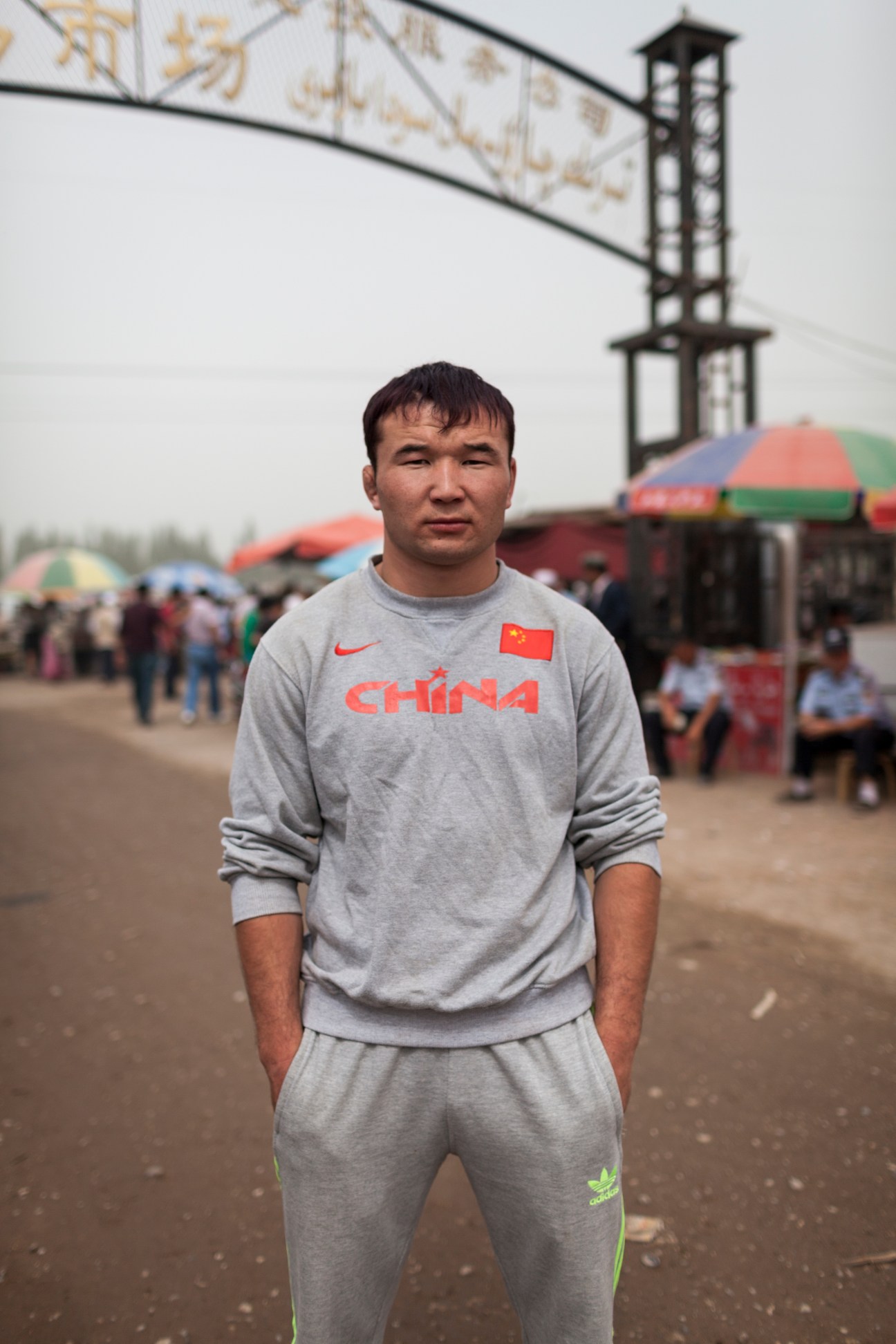 Man in grey China tracksuit stands at Kashgar Livestock Market with colorful umbrellas and mosque minaret behind.