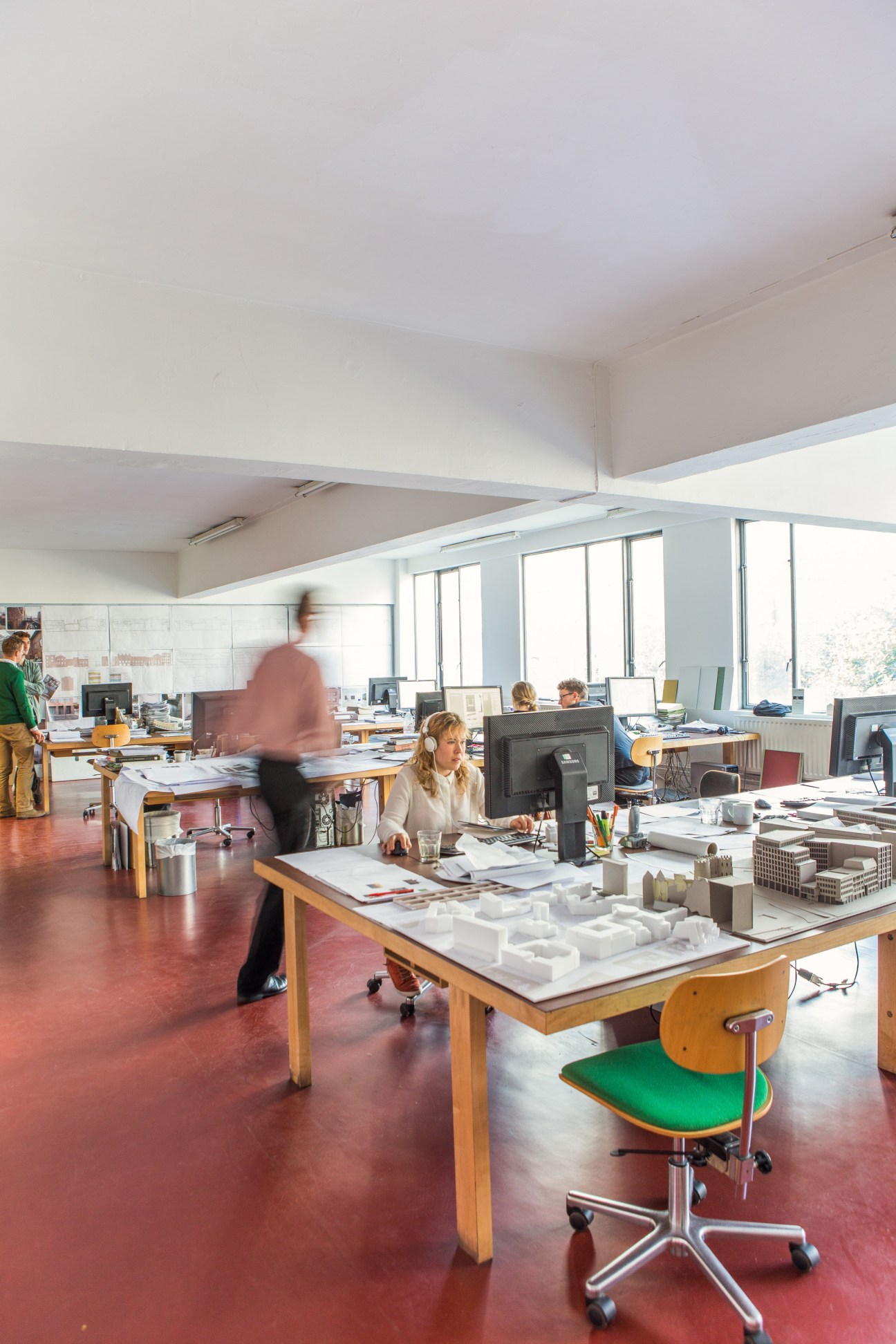Architects working at wooden desks in a bright, open studio space with red flooring and large windows.