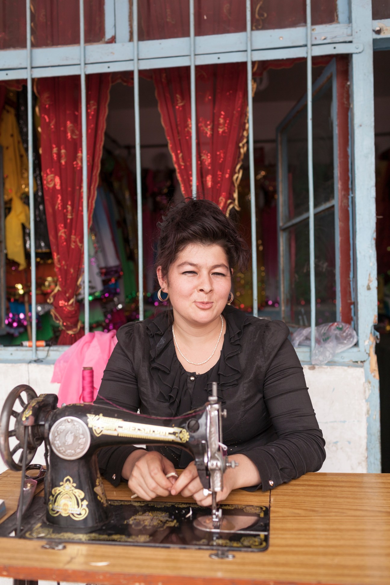 Seamstress at Kashgar Sunday Bazaar sitting at a sewing machine with red curtains behind her.