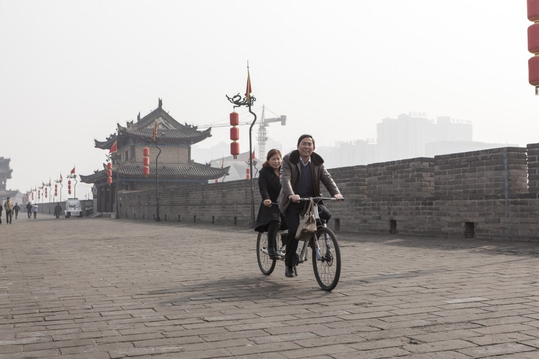 Two people riding a bicycle on a paved plaza beside Xi'an's ancient fortified city wall with traditional pagoda structures and red lanterns visible in the background.