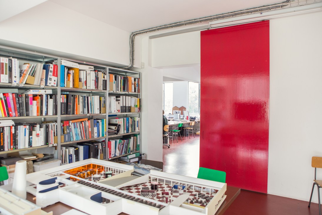 Interior of Caruso St John Architects' office in a converted factory with bookshelves, architectural models, and a bold red partition wall.