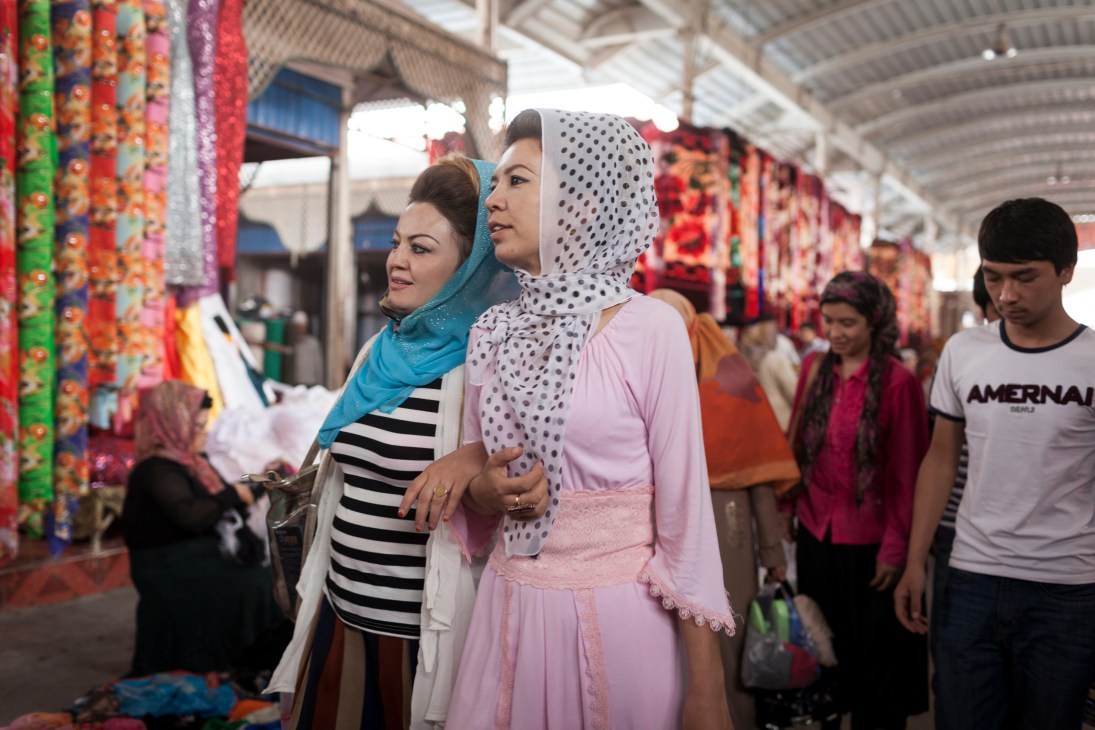 Women in headscarves shopping at Kashgar Sunday Bazaar among colorful fabric stalls.