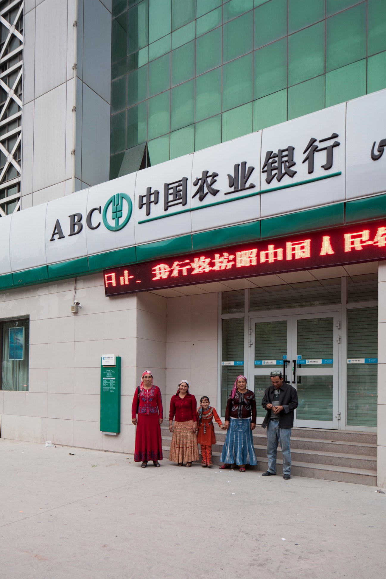 Uyghur family standing outside a modern bank building in downtown Kashgar with Chinese signage.