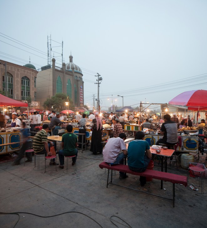Kashgar Night Market with diners at tables, food stalls under red umbrellas, and a mosque with green dome in the background.