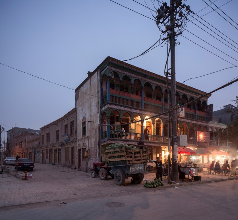 Traditional ornate building with arched windows at dusk in Kashgar's old town, with a loaded truck and vendors visible on the street.