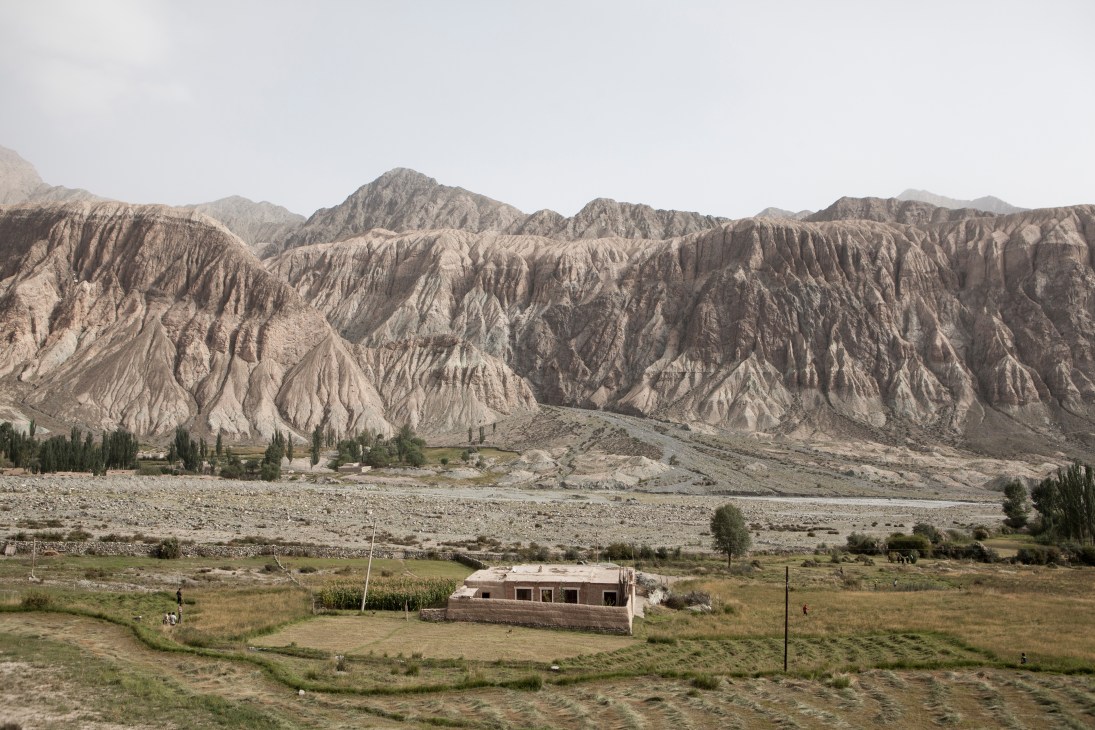 Arid mountain landscape with eroded ridges and a small brick building in a green valley below.