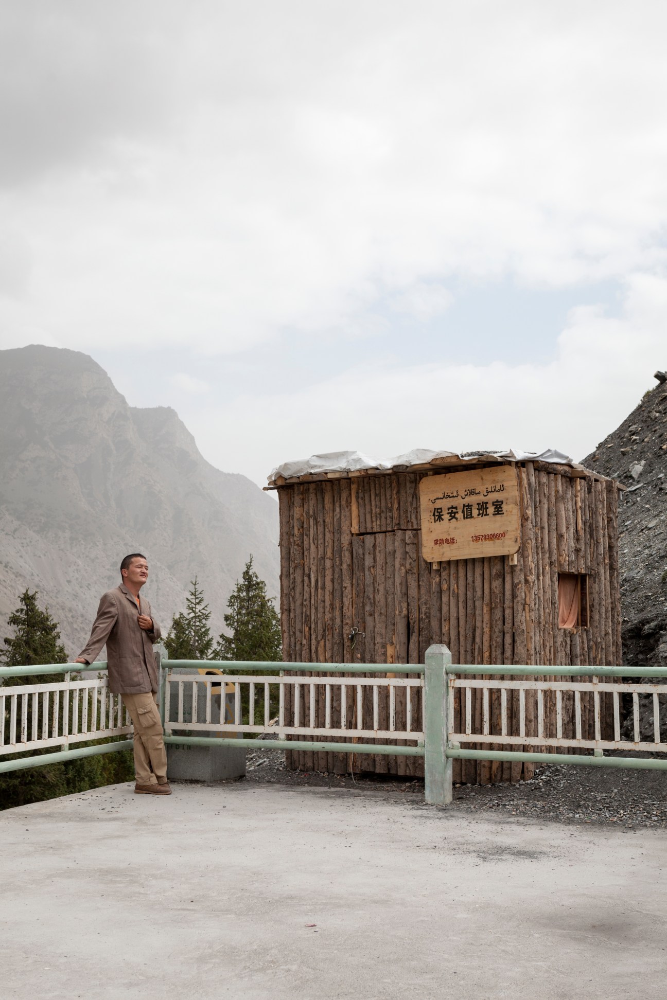 Local guide Mirsali Abudiwili stands at a viewing platform overlooking the Kizilsu Glacier, surrounded by mountains and evergreen trees.