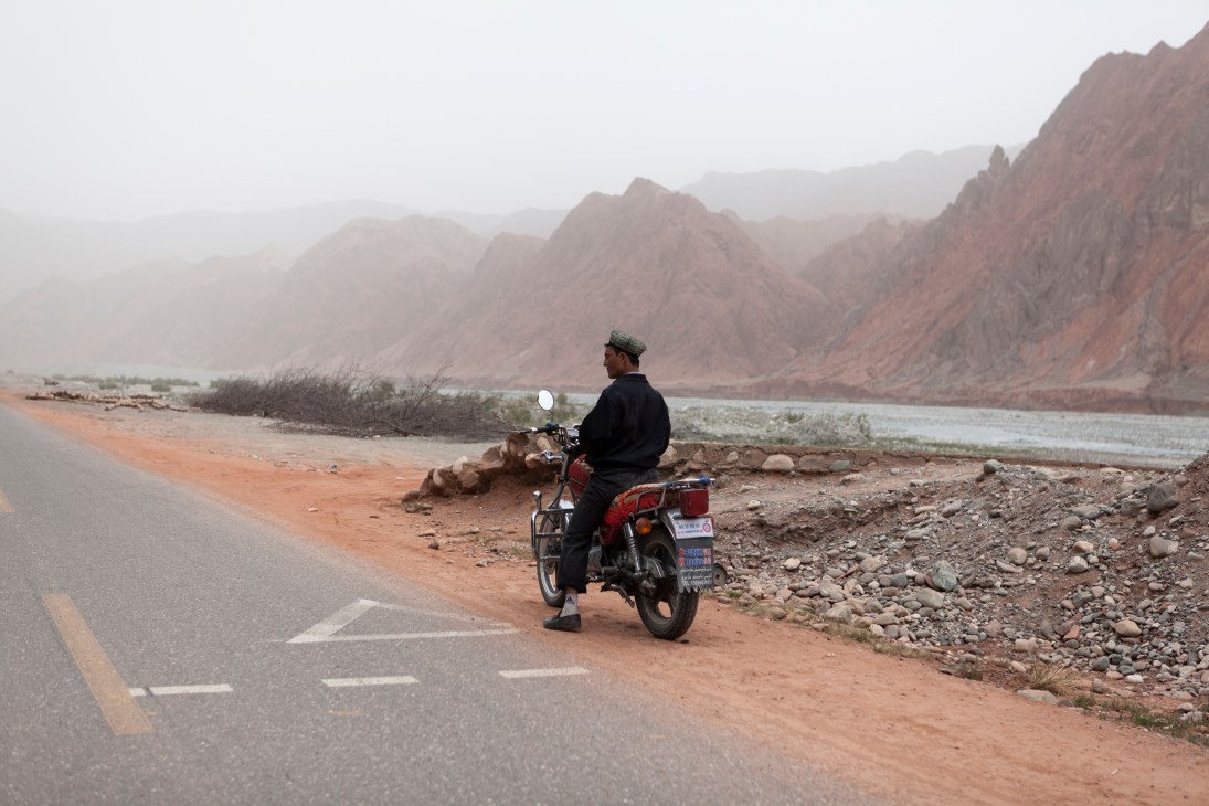 Man on red motorcycle taxi parked on Karakoram Highway with mountain river landscape.