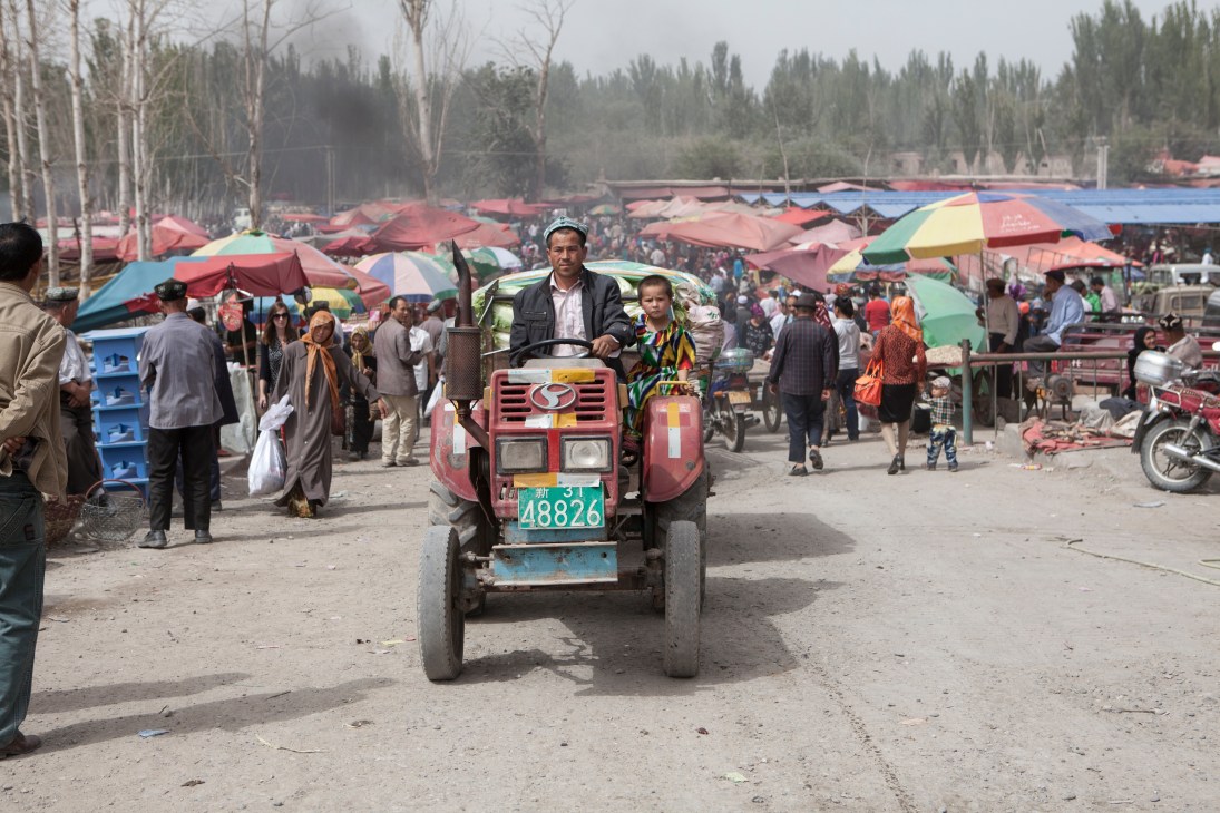 Man and child ride on a red tractor with sidecar through a busy market with colorful umbrellas.