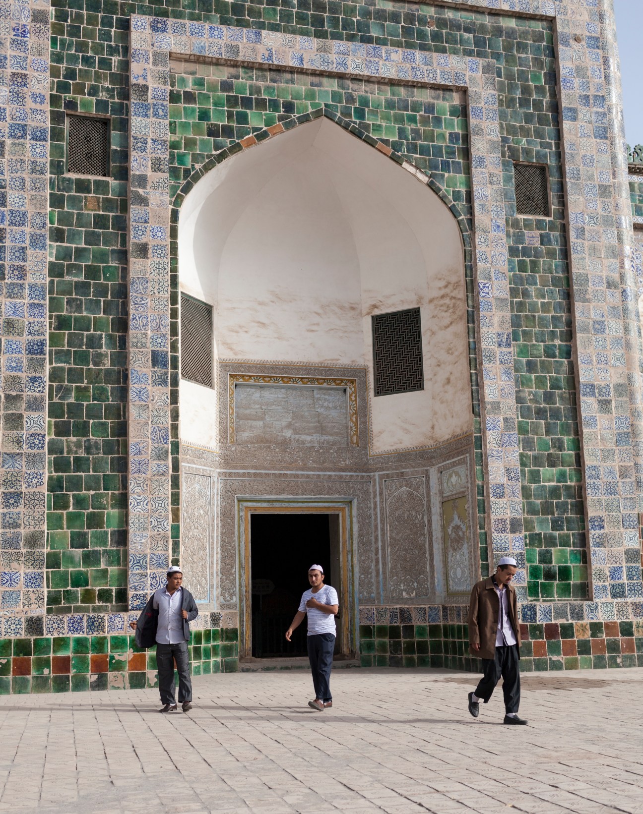 Three men stand in front of the Afaq Khoja Mausoleum with its distinctive green and blue tiled archway and intricate Islamic tilework.