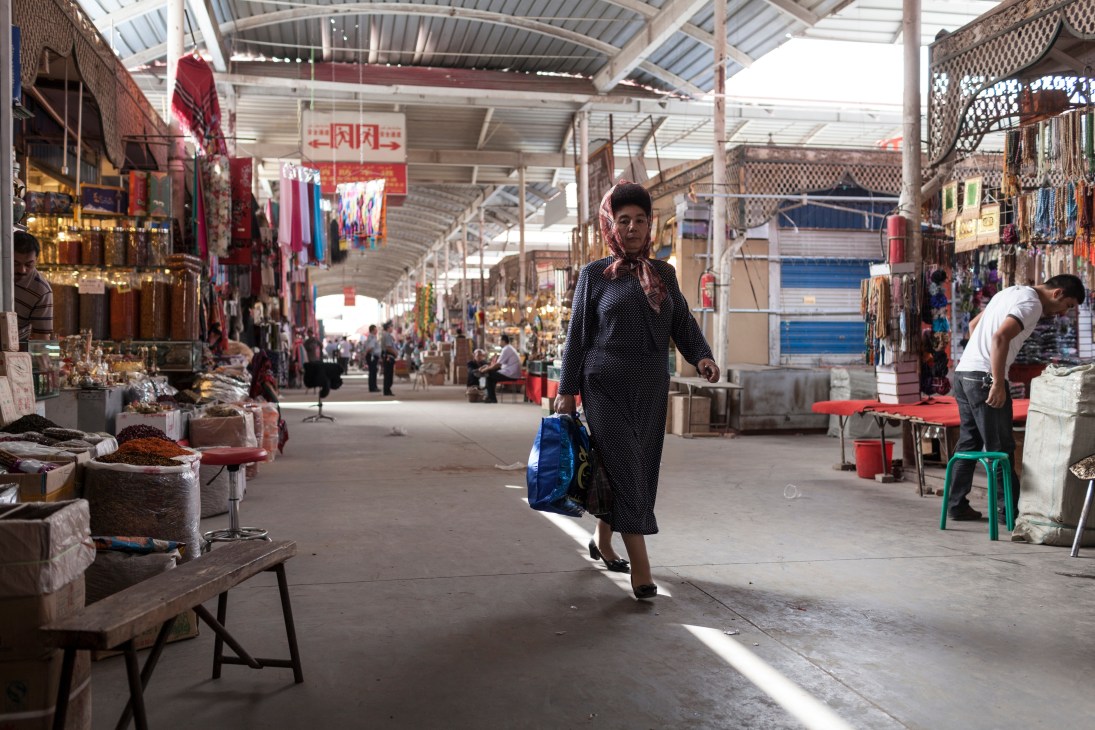 Woman in black polka dot dress carrying blue bag walks through Kashgar Sunday Bazaar amid vendor stalls.