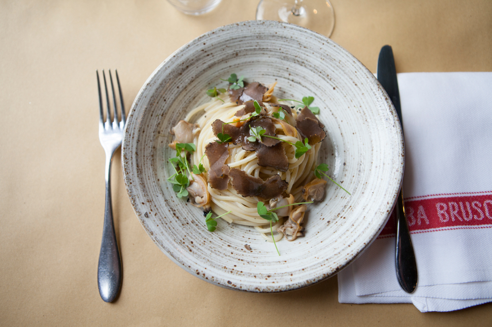 Spaghetti con vongole e tartufo plated in a speckled white bowl with clams, truffles, sorrel and parsley garnish.