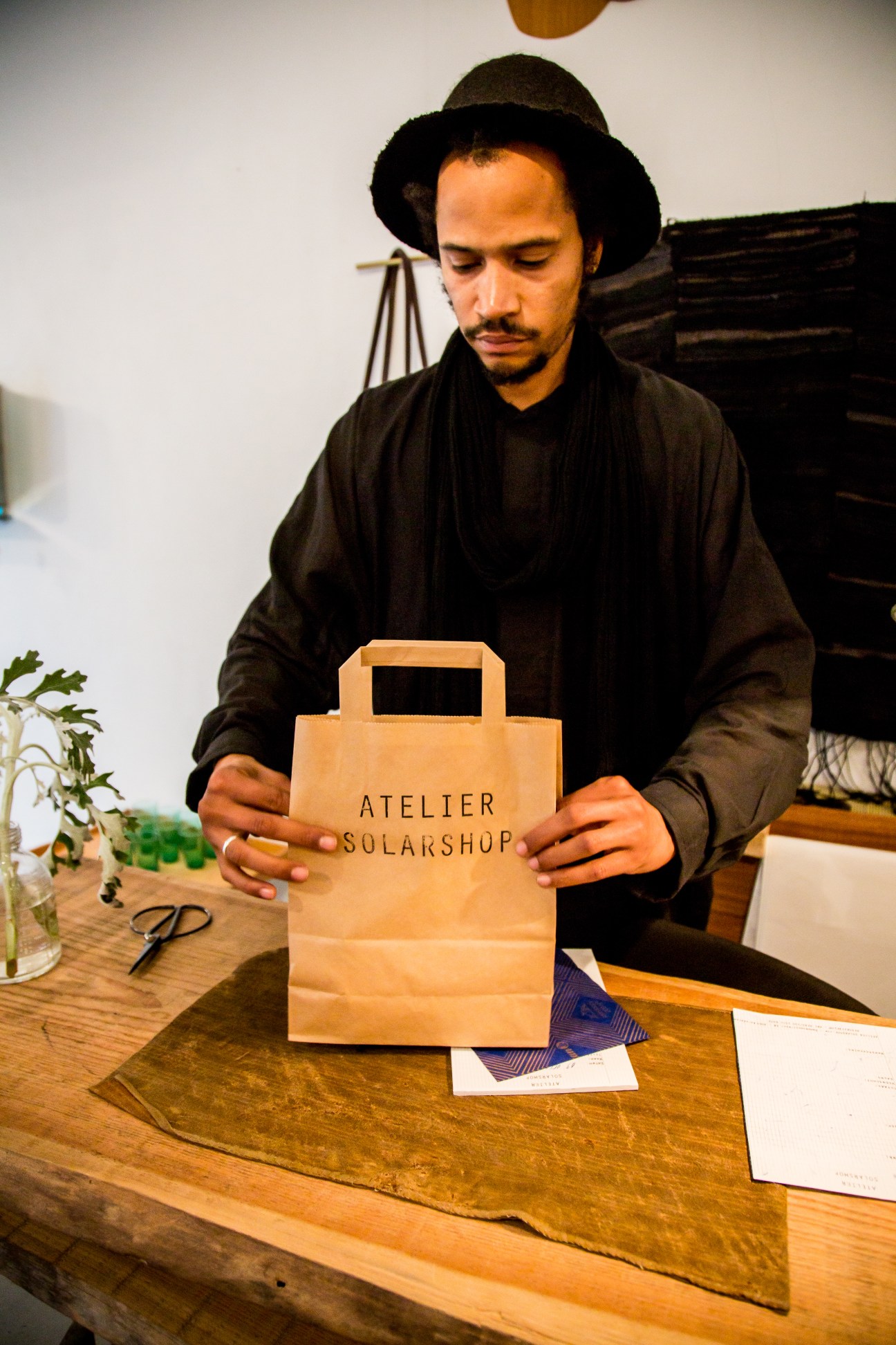 Man in black clothing and hat holds Atelier Solarshop paper bag at wooden table.