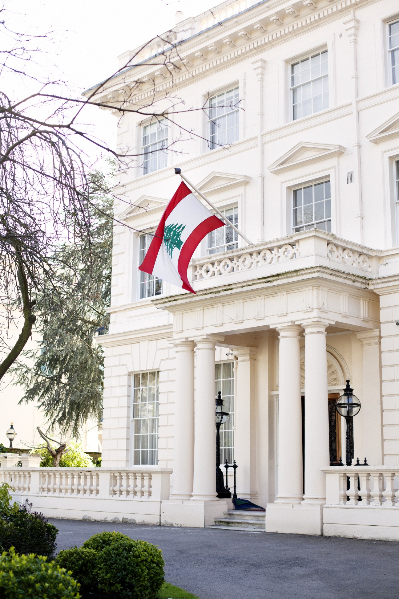 Lebanese flag flying outside the white stucco Embassy of Lebanon building in London, with classical columns and portico.