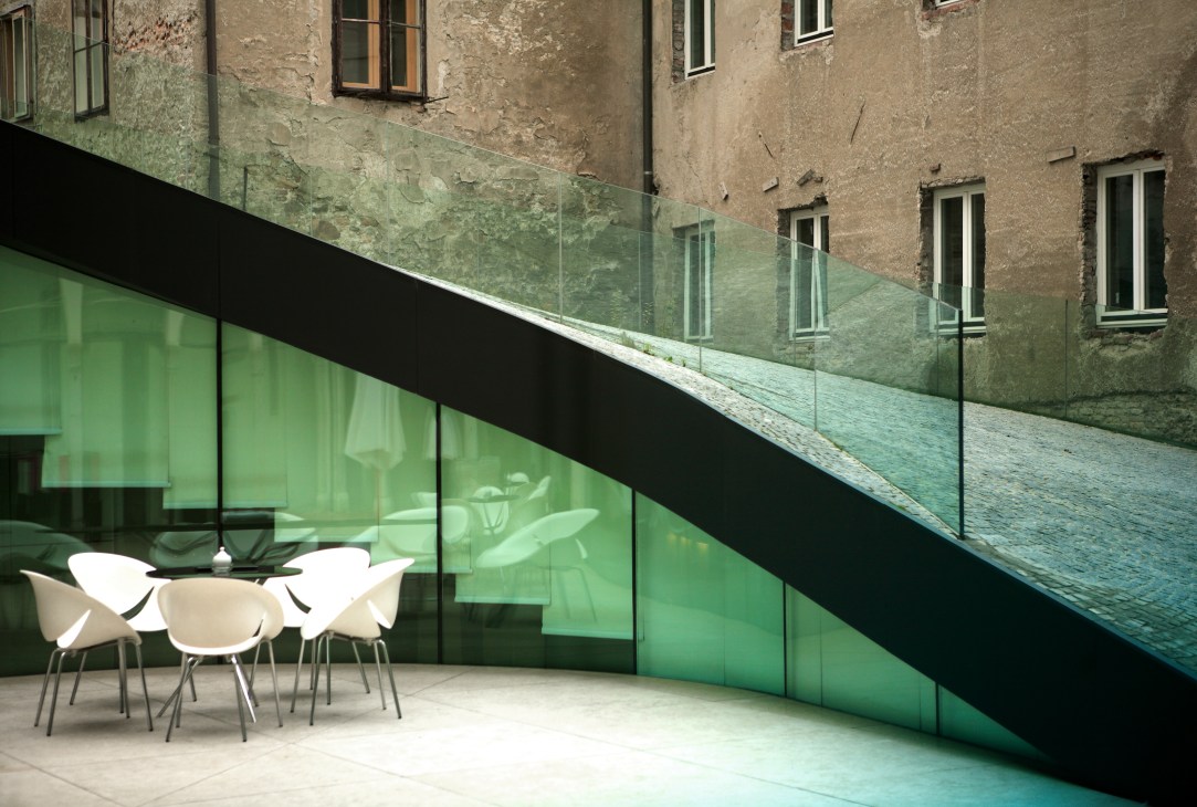 Modern glass and green paneled extension with outdoor seating attached to a historic beige stone building in Ljubljana.