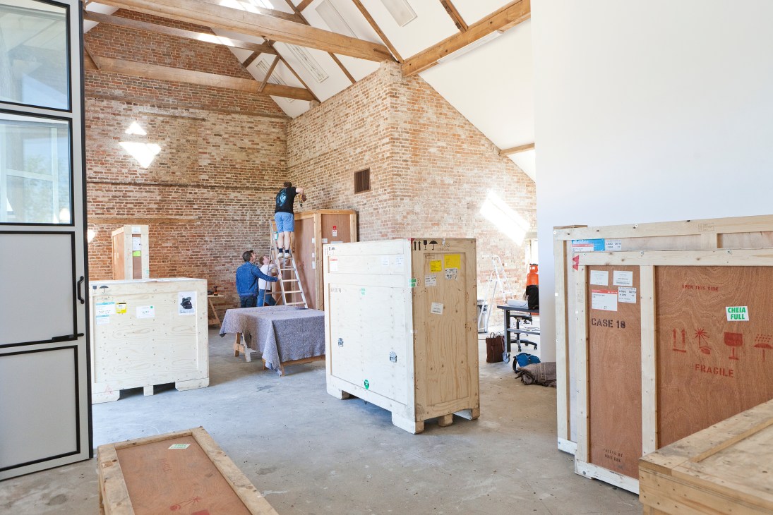 Technicians inspect and prepare wooden crates containing artworks in a converted barn with exposed brick walls and wooden beams.