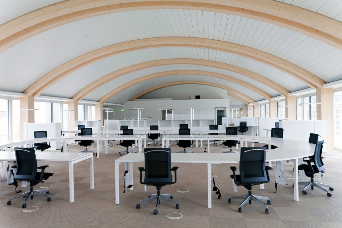 Circular open-plan news desk on seventh floor with wooden barrel-vault ceiling, white desks, and black office chairs.