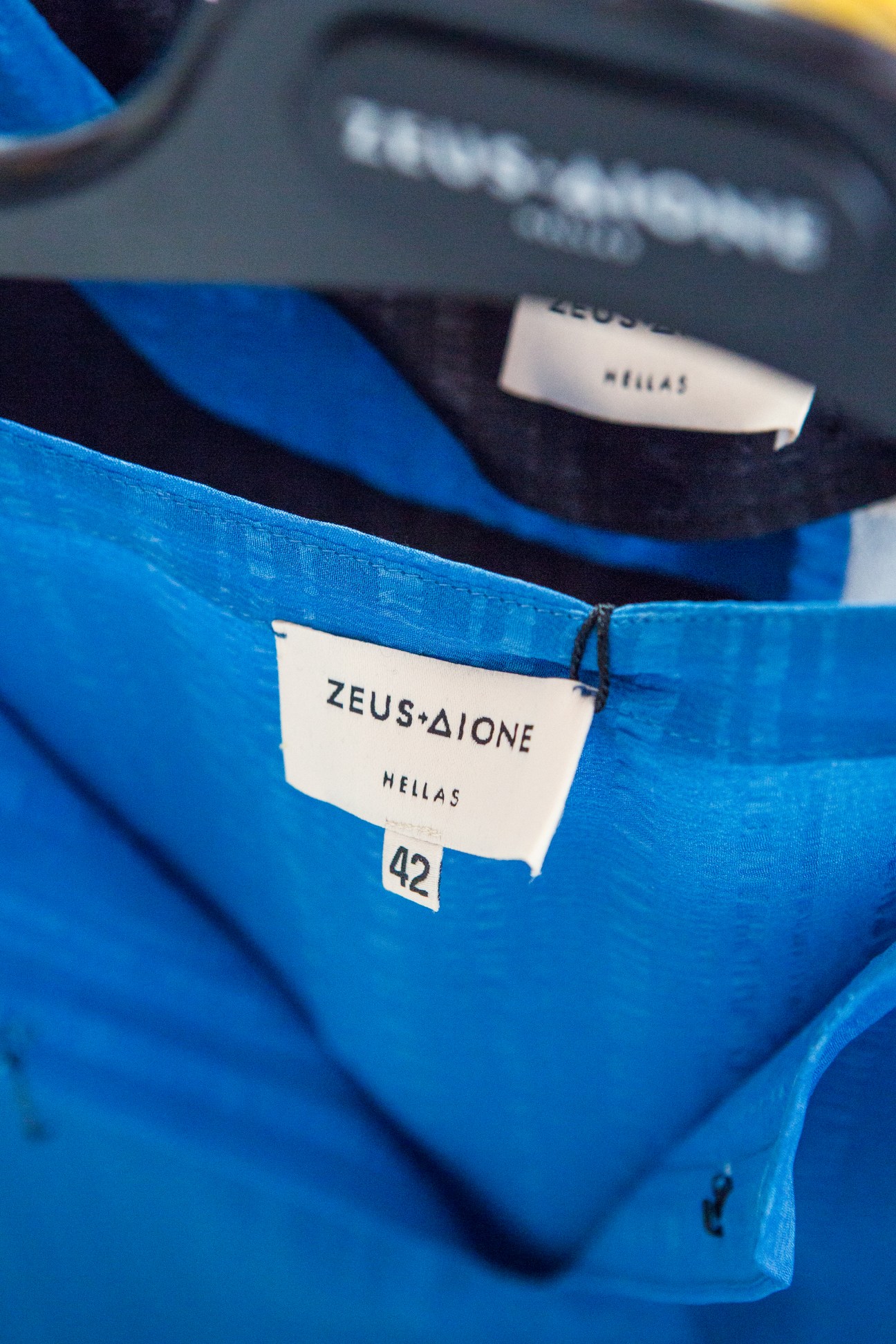 Close-up of Zeus + Dione garment labels on blue silk fabric showing brand name and Hellas origin.