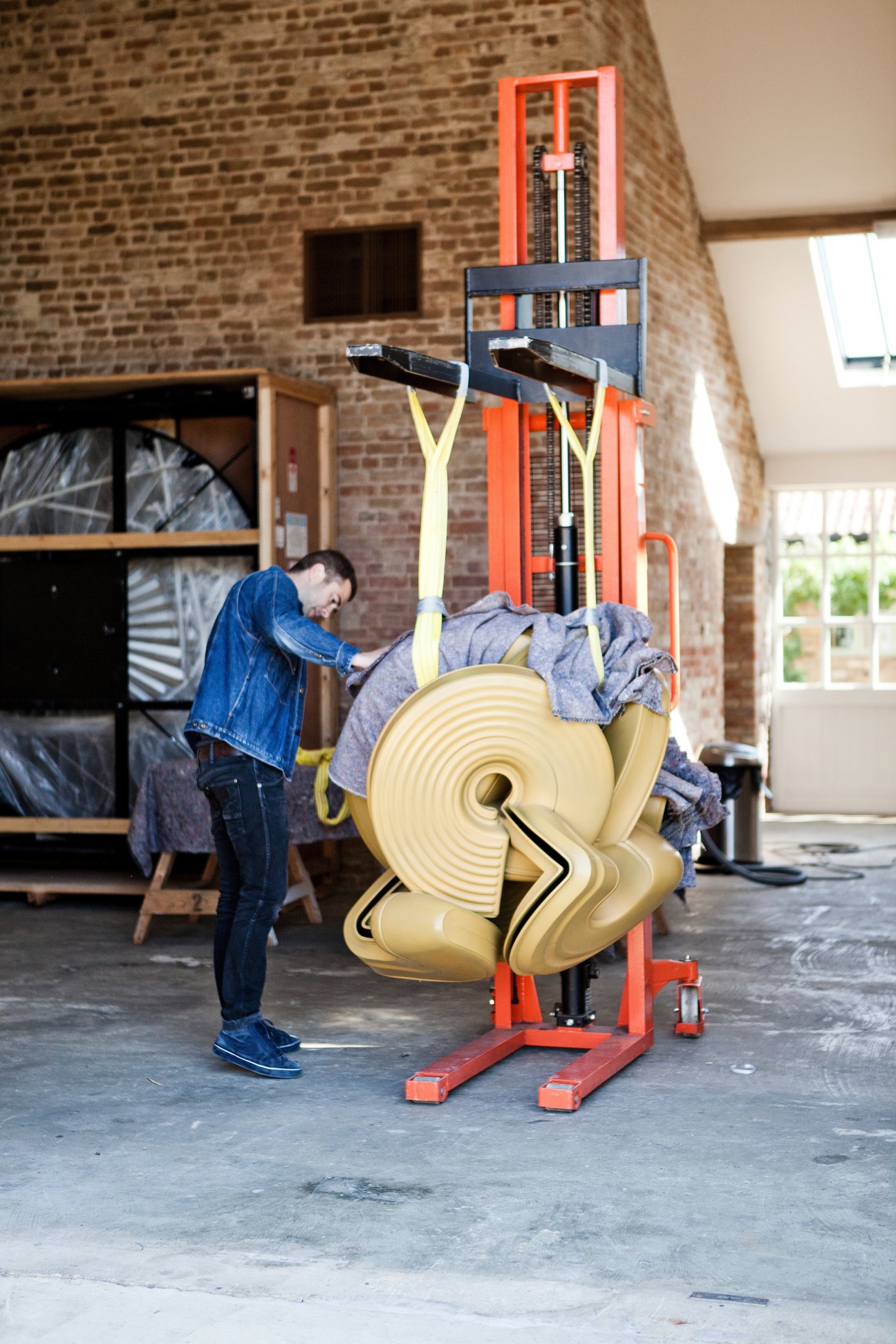 Technician adjusting a bronze sculptural artwork by Tony Cragg on a red industrial lift in a warehouse.