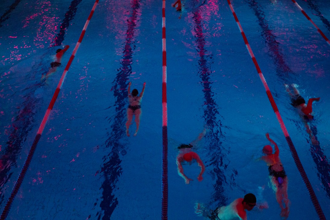 Overhead view of swimmers doing laps in a floodlit pool separated by red lane dividers.