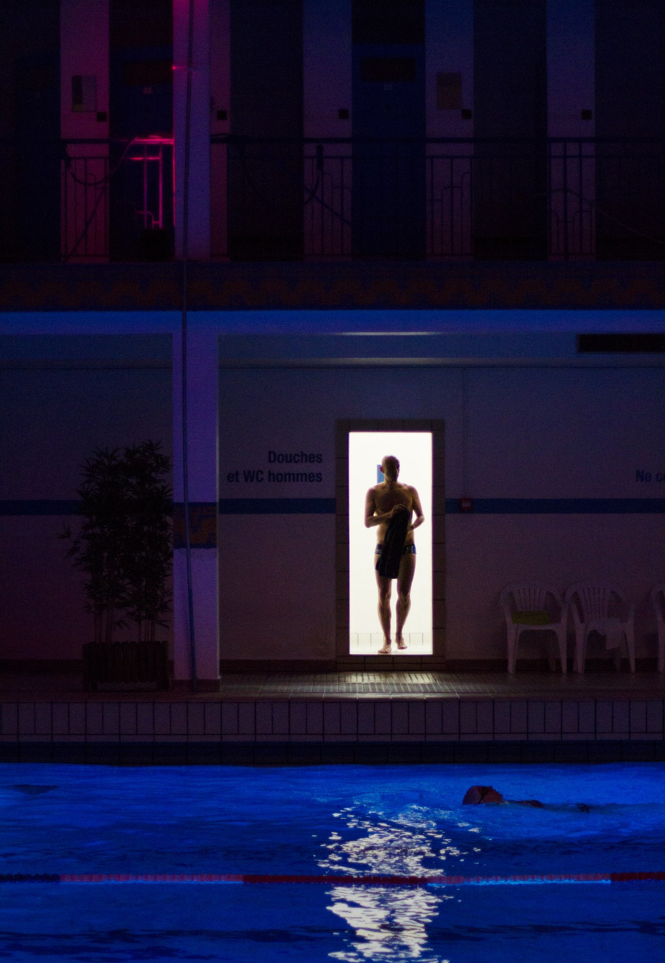 A swimmer in the brightly lit doorway of La Piscine de Pontoise at night, overlooking the illuminated blue pool below.