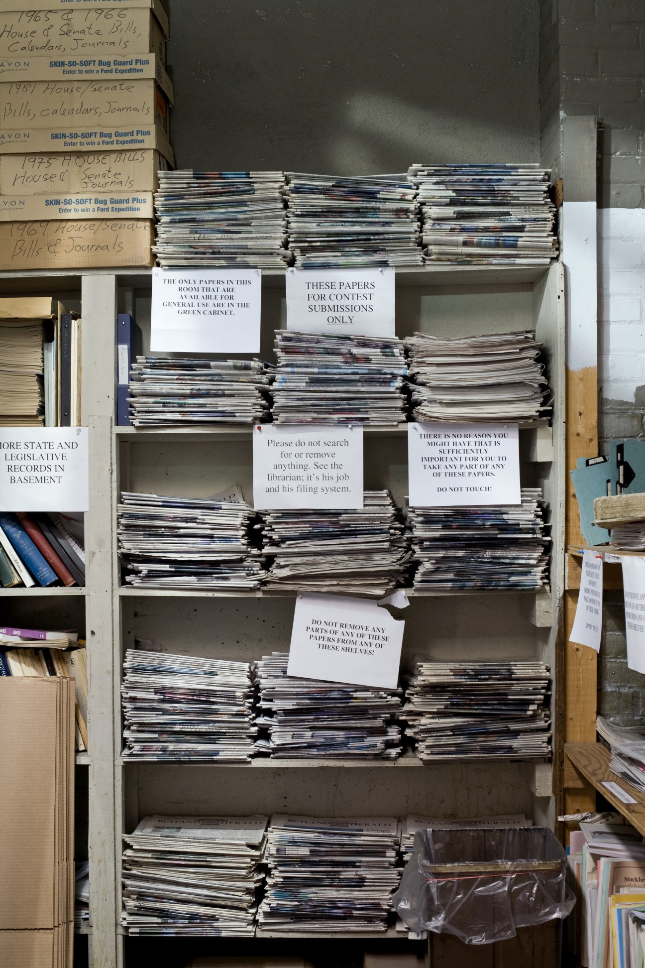 Metal shelving unit filled with stacked newspapers and archived documents, labeled with handwritten notes and instruction signs.