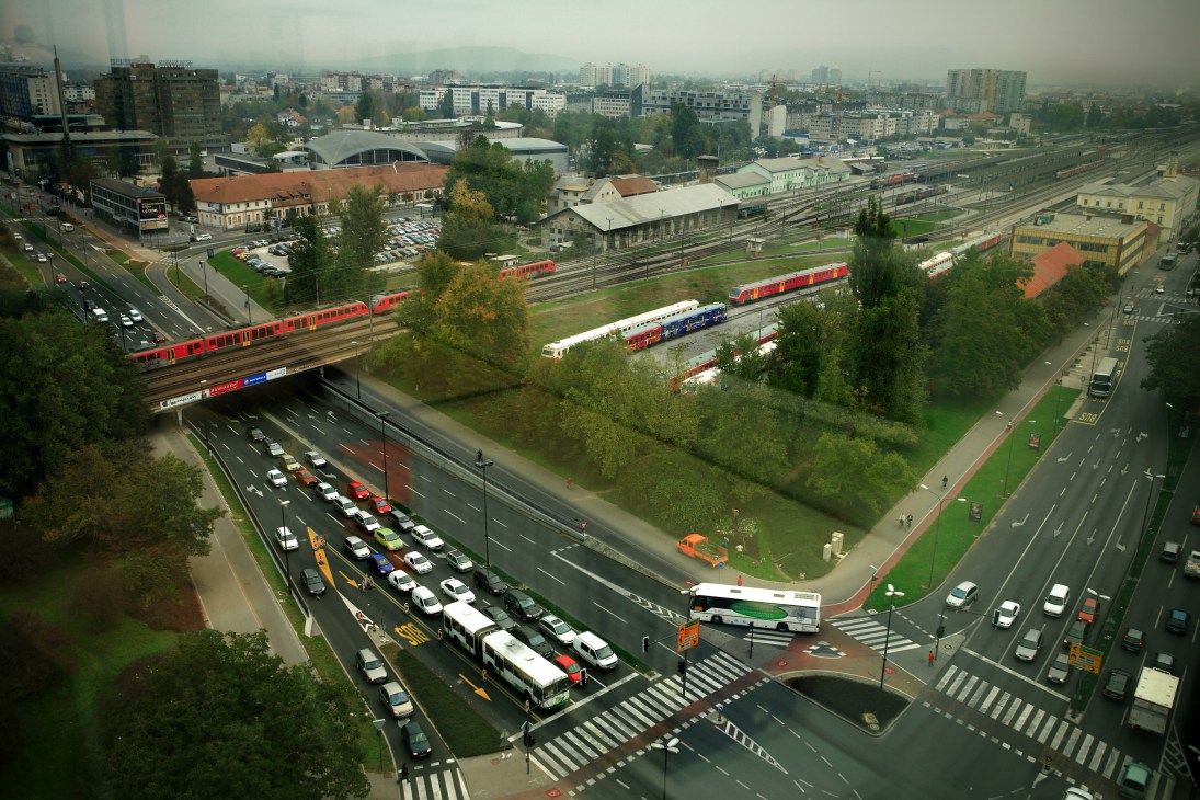 Aerial view of Ljubljana showing busy multi-lane arterial roads with trains, cars, and urban buildings beneath overcast sky