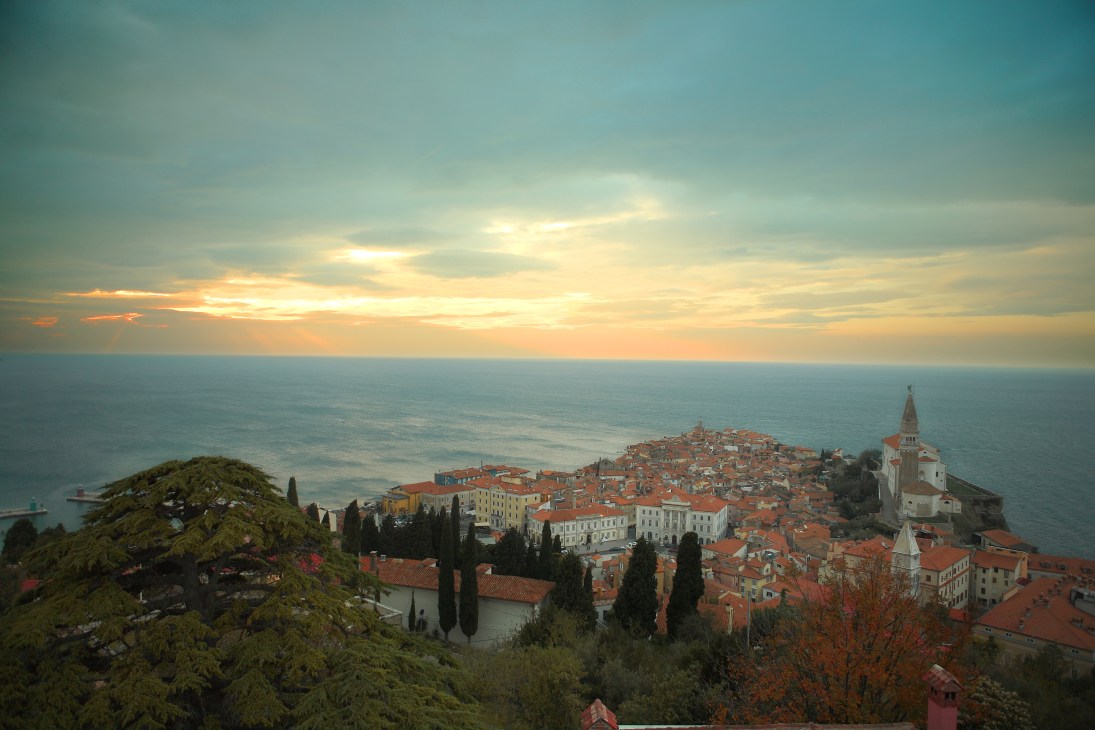Coastal town of Piran with red-roofed buildings, church bell tower, and Adriatic Sea at sunset.