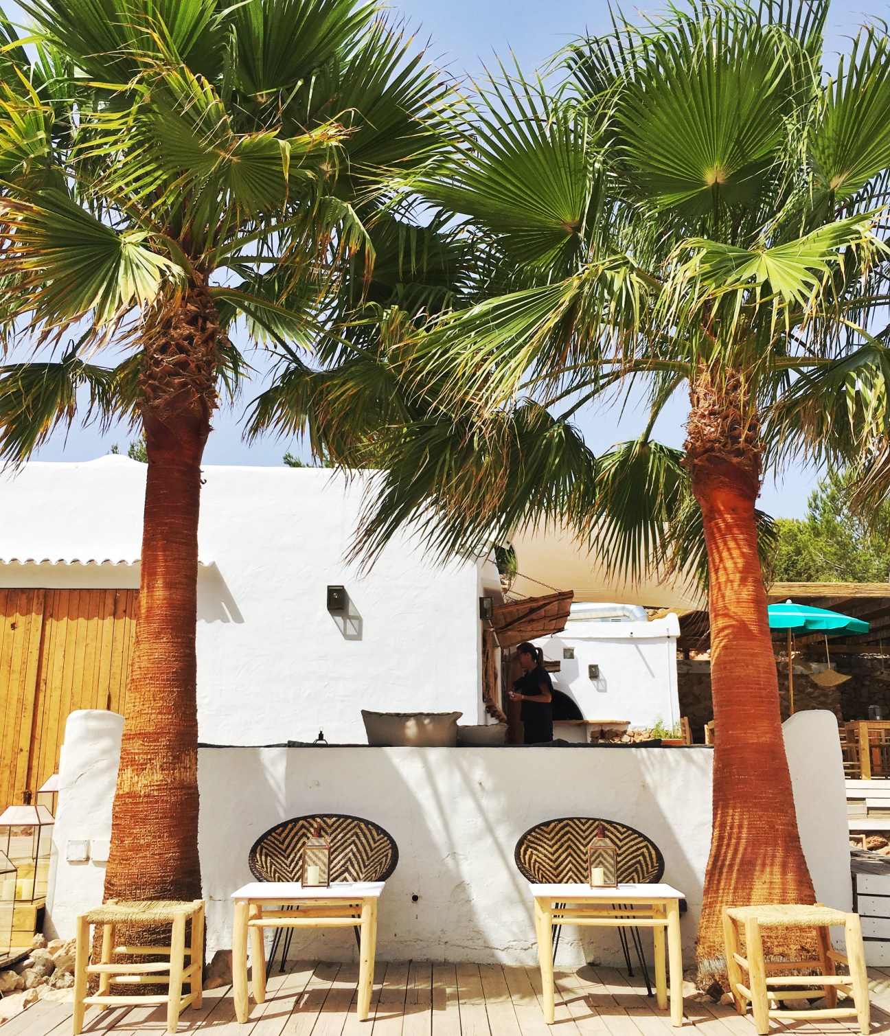 Beach bar seating area with palm trees, white walls, wooden chairs, and turquoise umbrella at Cala Bonita, Ibiza.