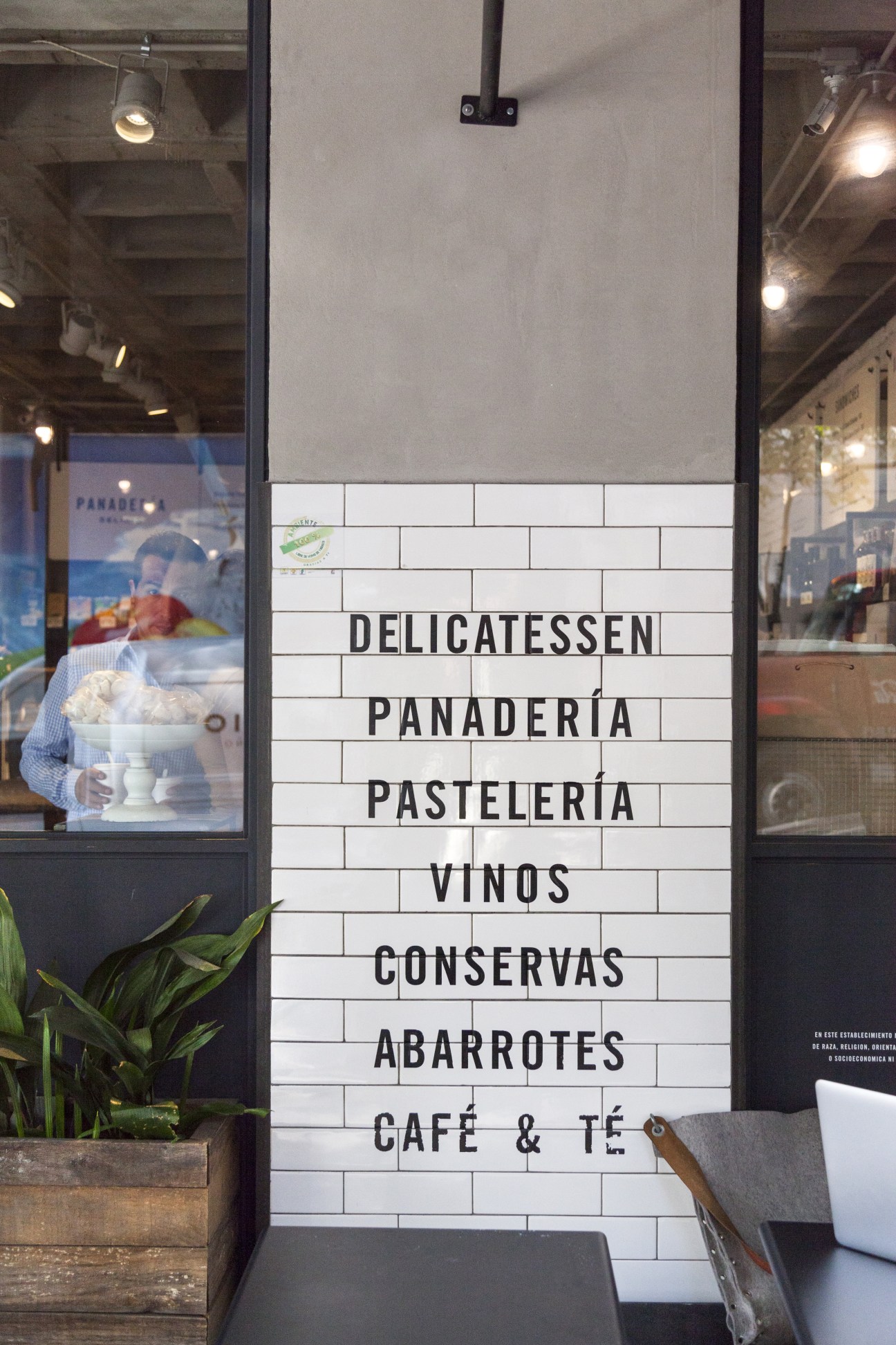 Abarrotes storefront window displaying white tiles with black letters listing offerings including delicatessen, bakery, pastries, wines, preserves, groceries, and café.