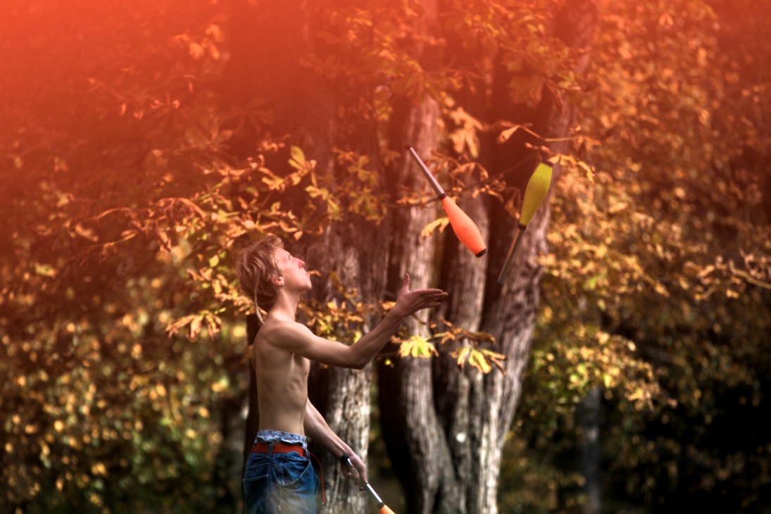 Shirtless juggler tosses clubs in Tivoli Park, Ljubljana, surrounded by autumn foliage.