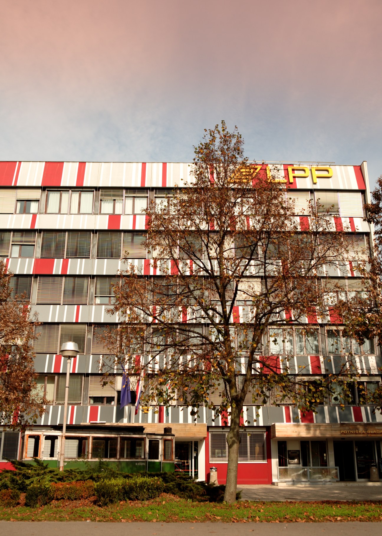 Red and white striped modernist office building in Ljubljana with bare tree in front.