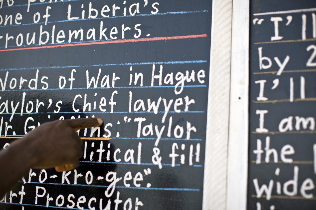 A hand points to handwritten chalk headlines on a blackboard newspaper, including stories about Liberia's troublemakers and war in Hague.