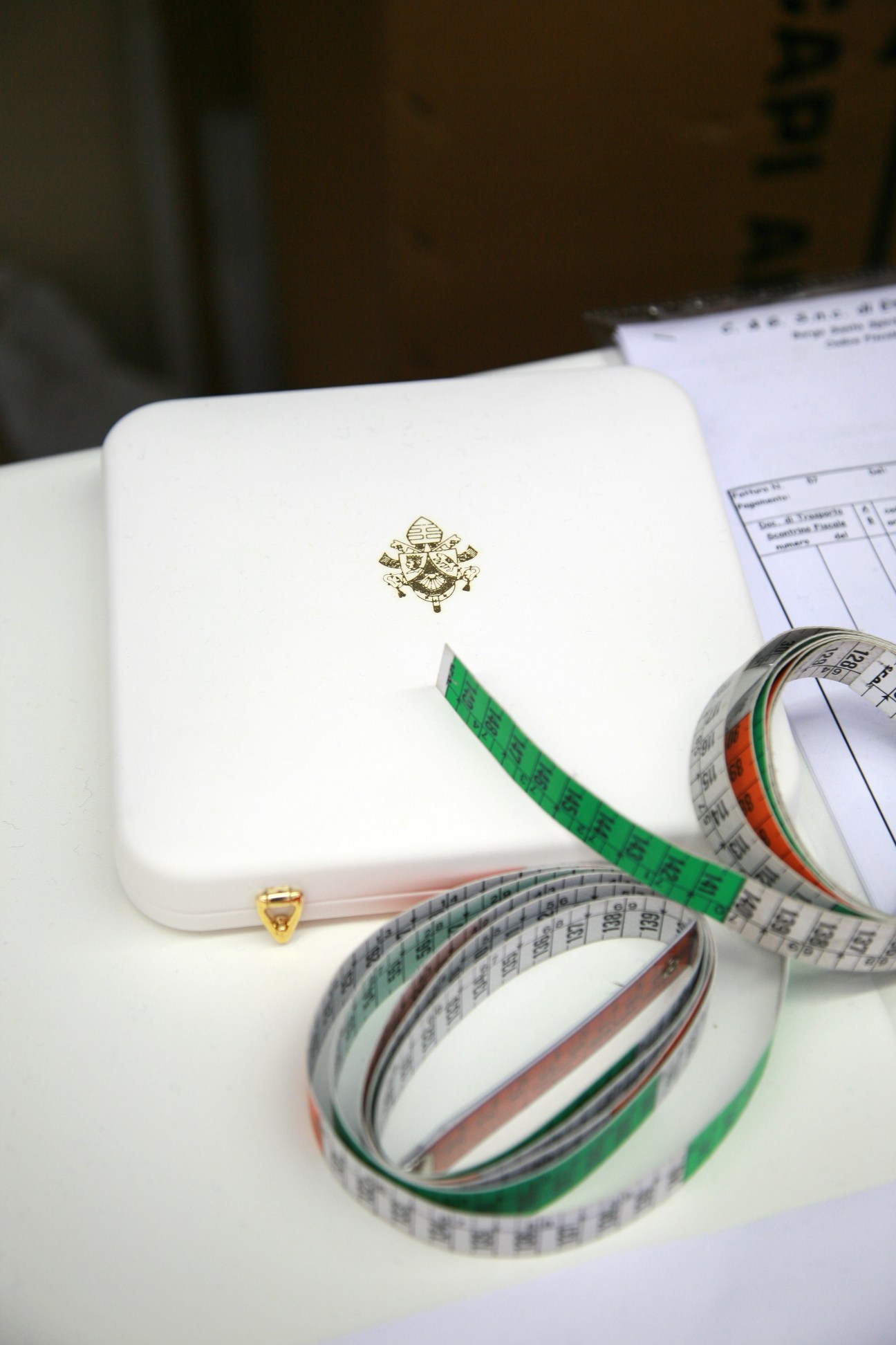 Italian flag measuring tape coiled next to a white case with papal emblem on tailor's desk.