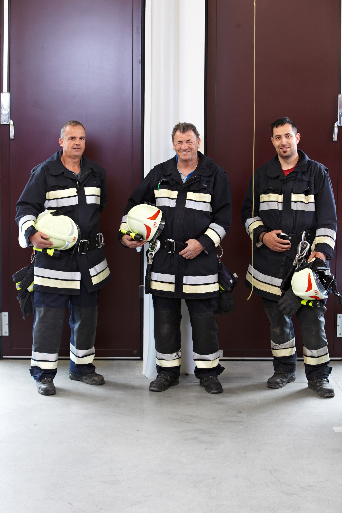 Three firefighters in black uniforms with reflective stripes holding white helmets stand in front of a purple wall.