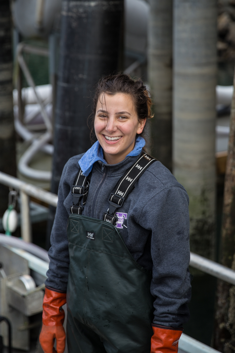 Abi Morris, deckhand, smiles at the camera wearing waterproof gear on a fishing dock.