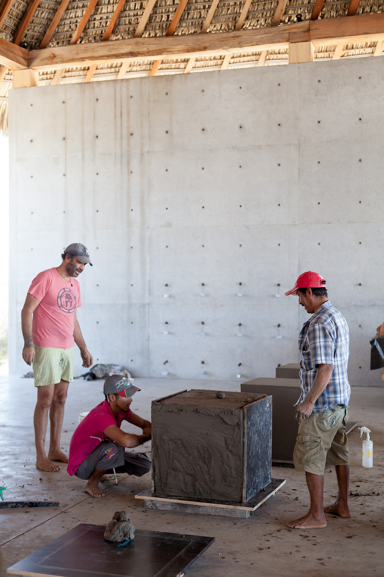 Bosco Sodi in a pink shirt and cap works with collaborators on large clay cubes under a wooden and thatched-roof structure.