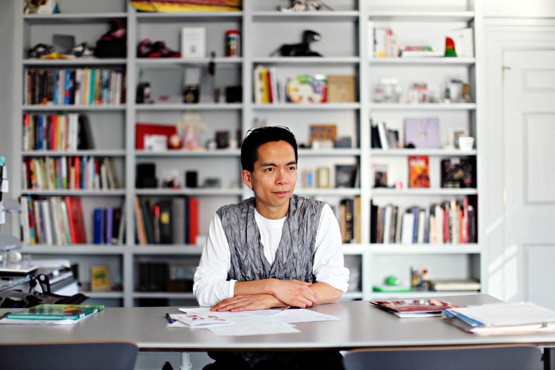 John Maeda sits at a desk in his office with white shelving filled with books and design objects behind him.