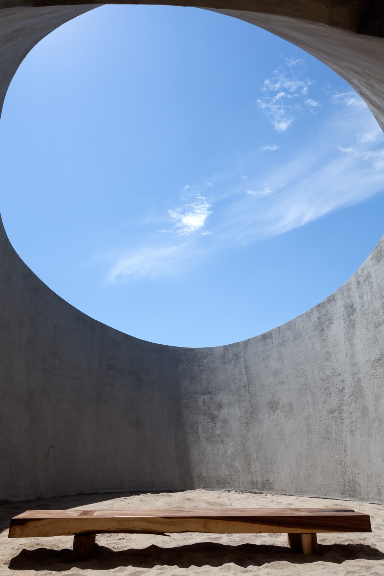 A circular opening in a concrete wall frames a bright blue sky with wispy clouds, with a wooden bench visible below.