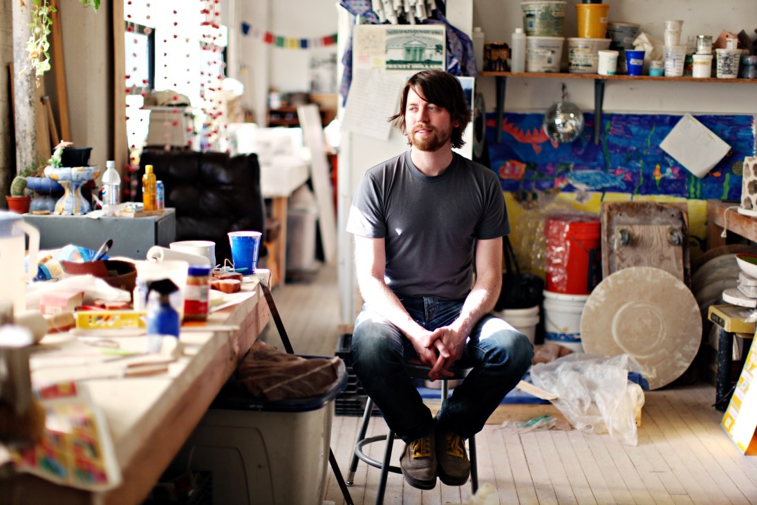 Noel O'Connell, a fine art ceramics graduate, sits in his studio surrounded by pottery wheels, ceramic pieces, and art supplies.